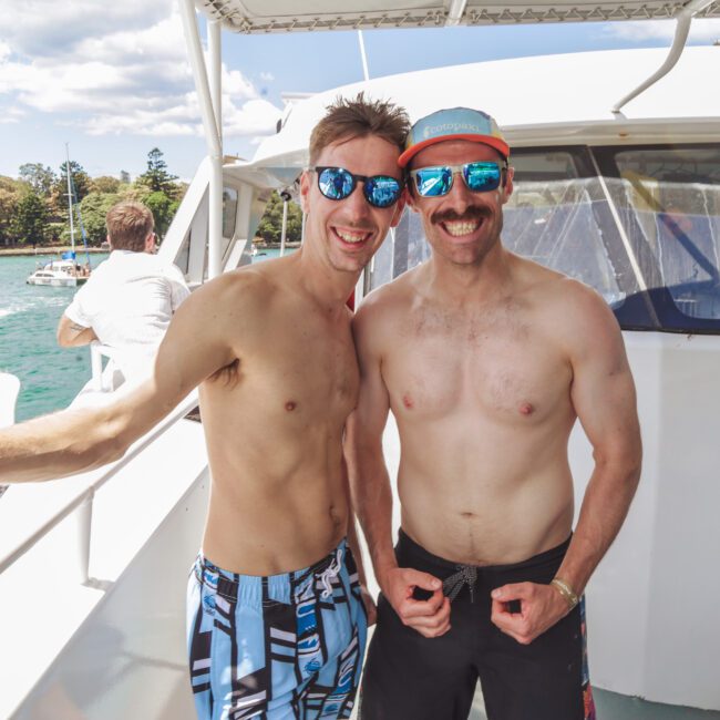 Two smiling men in swimwear and sunglasses pose on a boat under sunny skies, with water, trees, and another boat visible in the background.