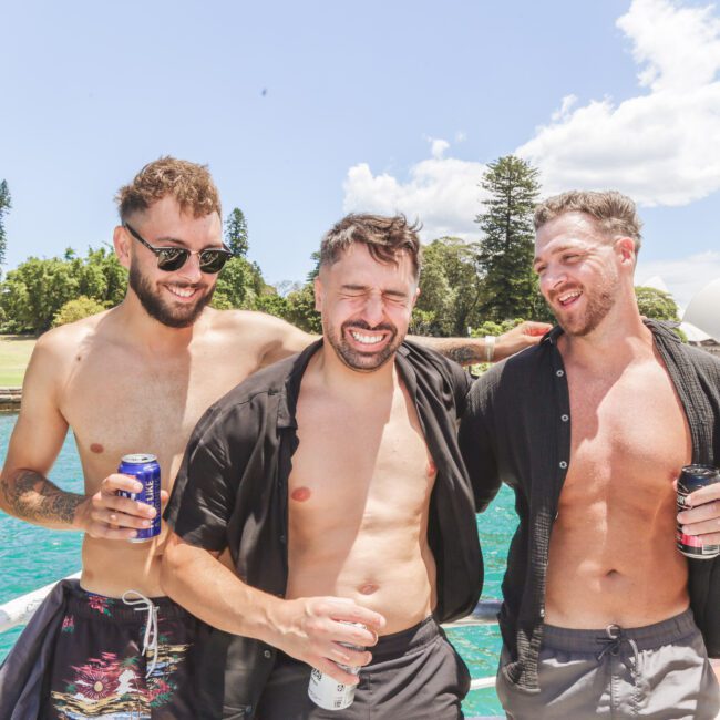 Three shirtless men stand smiling and laughing together on a boat with drinks in hand, with the Sydney Opera House, trees, and blue water in the background on a sunny day.