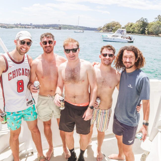 Five men stand together smiling on a boat in sunny weather, wearing casual summer clothes and holding drinks, with water, boats, and trees visible in the background.