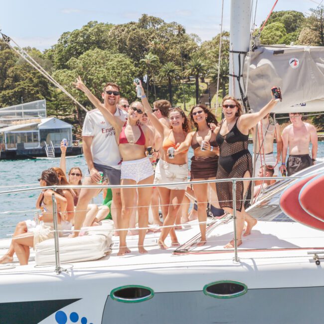 A group of people in swimsuits stand and pose together on the deck of a sailboat, smiling and holding drinks. The boat is on the water, and trees and buildings are visible in the background.