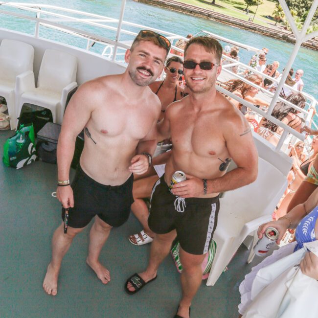 Two smiling, shirtless men in swim trunks stand on a boat deck, holding drinks; people sitting and enjoying the sunny weather are visible in the background with green trees and water nearby.