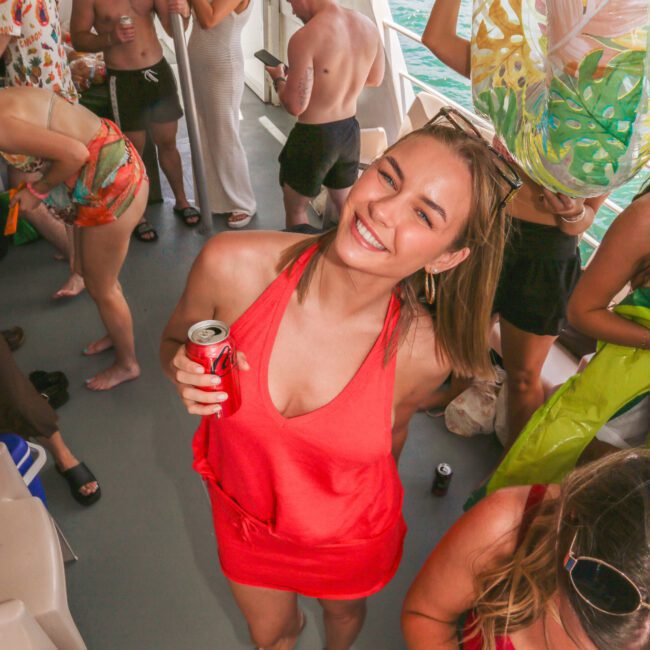 A woman in a red dress smiles at the camera holding a can of Coke on a boat, surrounded by casually dressed people socializing and enjoying drinks near the water.