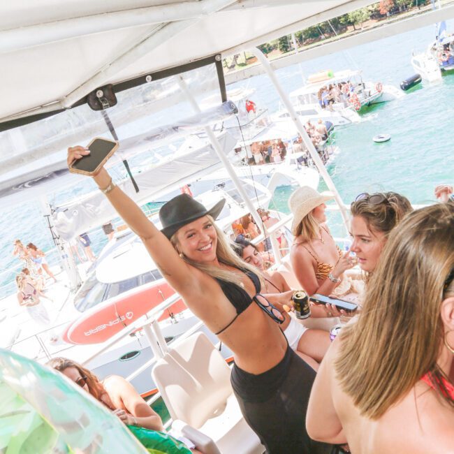 A woman in a black bikini and hat poses with her arm raised and smiling on a crowded boat with other people. The boat is decorated with green leaves and is surrounded by water and other boats under sunny skies.