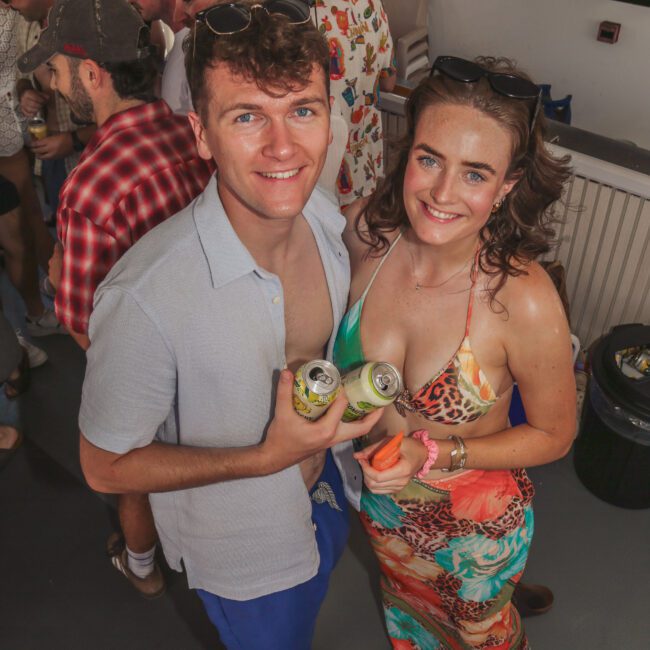 A man and woman smiling at a crowded indoor party, holding cans. The woman wears a colorful dress and has a prosthetic leg. They are looking up at the camera, surrounded by other casually dressed people.