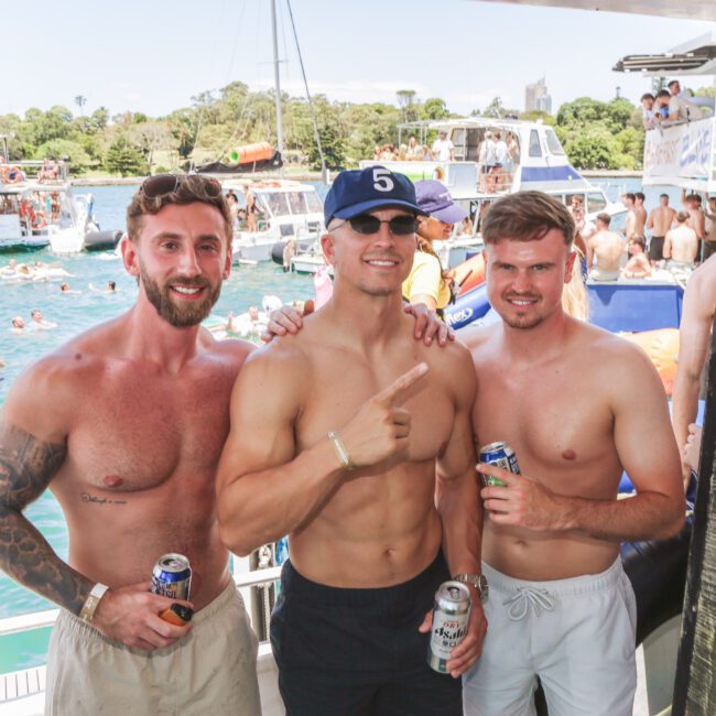 Three shirtless men posing and smiling on a boat, holding drinks. In the background, people are swimming and partying on nearby boats, with trees and blue water visible. It's a sunny day and a lively atmosphere.