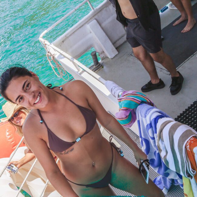 A woman in a brown bikini smiles at the camera on a boat, with colorful towels on the railing and water in the background. Another person in a hat sits near the edge, and others stand nearby.