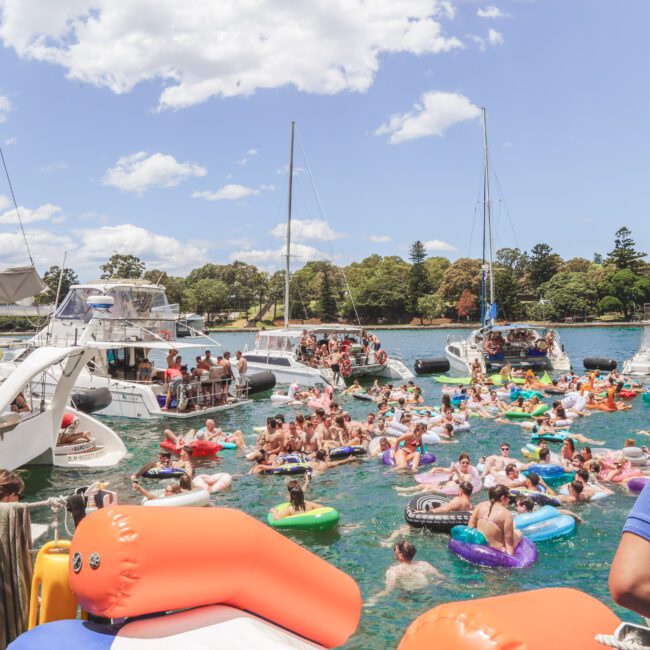 A lively scene of people on colorful pool floats and inflatables in the water, surrounded by several anchored boats. Crew members stand on a boat, overseeing the fun under a sunny sky with scattered clouds.