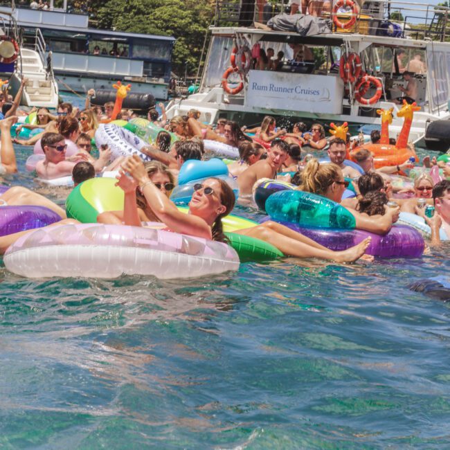 A large group of people relax and socialize on colorful pool floats in the water near several boats on a sunny day. Some people are sunbathing, and others are chatting, with trees and blue sky in the background.