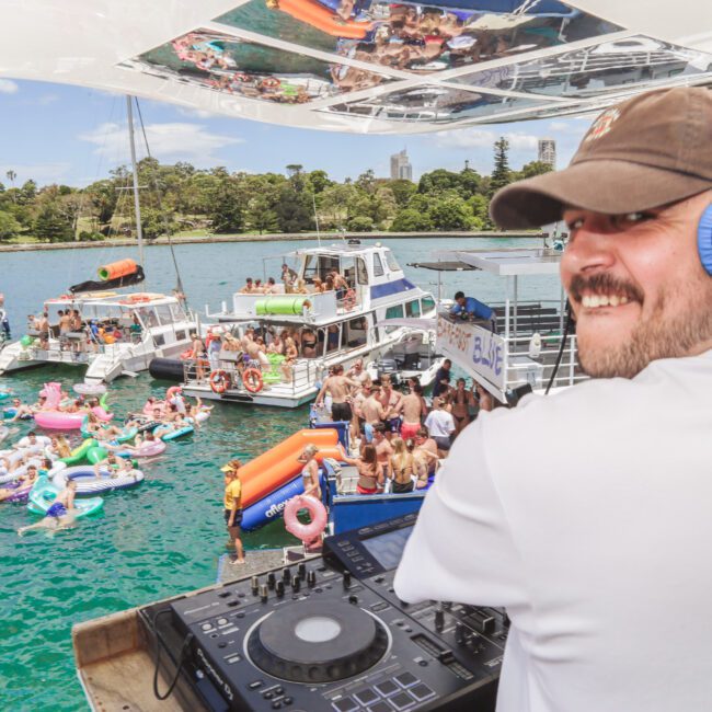 A DJ wearing headphones and a brown cap smiles at the camera while playing music on a boat, overlooking a lively crowd swimming and relaxing on inflatables near other boats on a sunny day.