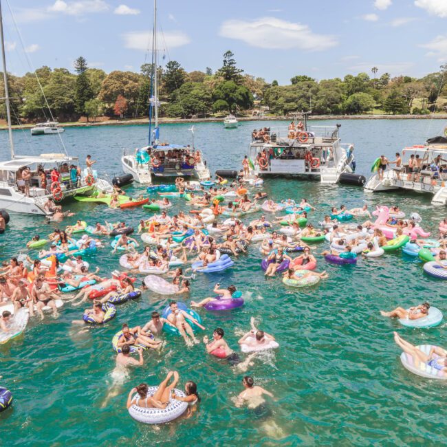 A large group of people enjoy a sunny day floating on colorful inflatables in the water, surrounded by several boats anchored nearby, with trees and blue sky in the background.