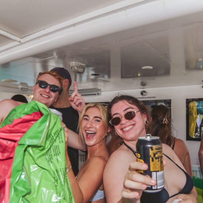 Three young adults smile and pose for the camera on a boat, holding drinks and an inflatable float. Others are in the background, suggesting a lively party atmosphere.