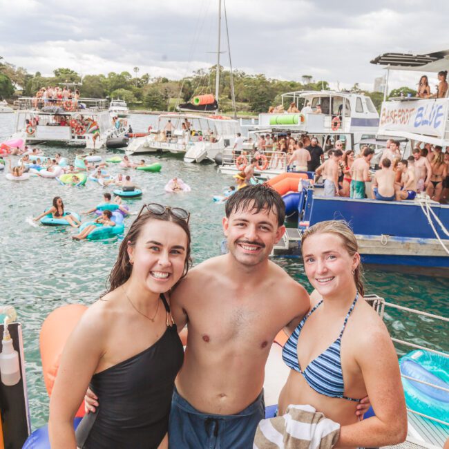 Three people in swimsuits smile for a photo at a lively boat party, with many others swimming, lounging on floats, and gathering on boats in the background on a sunny day.