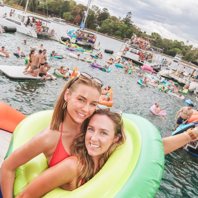 Two smiling young women pose on a boat; one wears a green inflatable ring around her. Behind them, many people enjoy swimming and relaxing on colorful inflatables in a lively river scene.