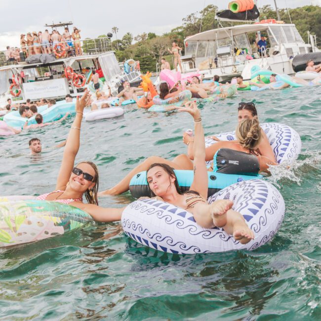 People relax on colorful inflatable tubes in the water near boats. Many are smiling, raising their arms, and enjoying a lively, social atmosphere under a cloudy sky. The scene feels festive and carefree.
