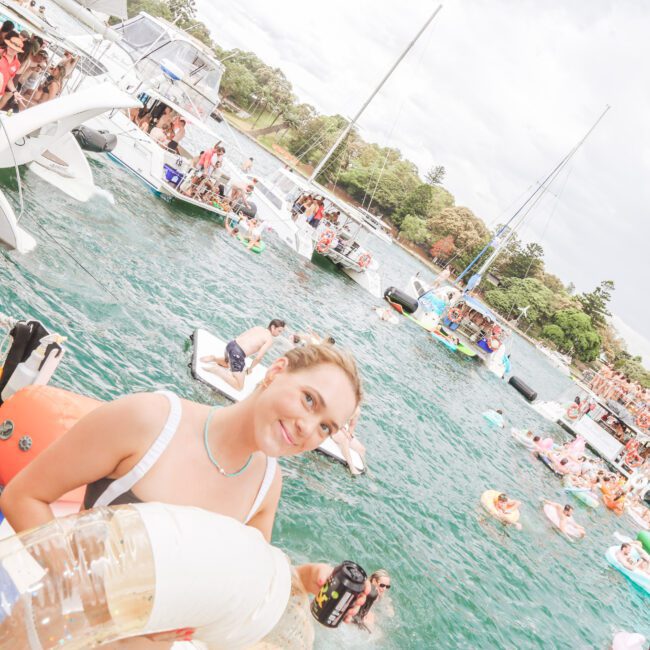 A young woman holding an inflatable ring stands near the edge of a crowded body of water with many people swimming, floating on inflatables, and boats anchored nearby during a lively outdoor event.