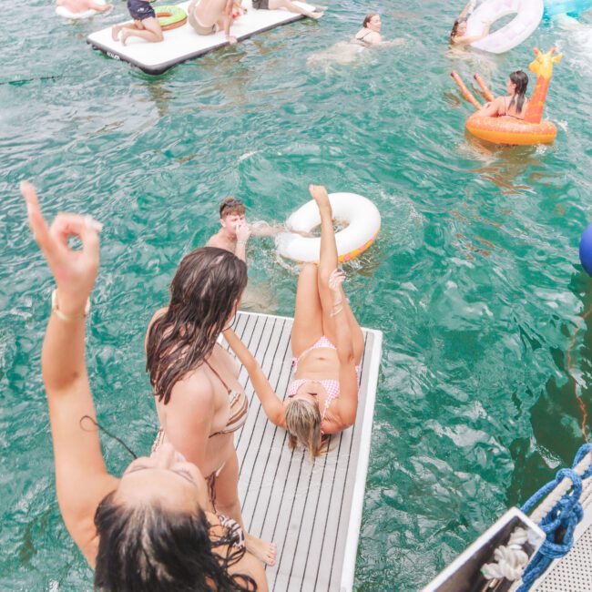 People in swimsuits enjoy a lively pool party with floaties and inflatables in the water; two women stand on a dock, one playfully pointing upward while another lounges on a float near the dock’s edge.
