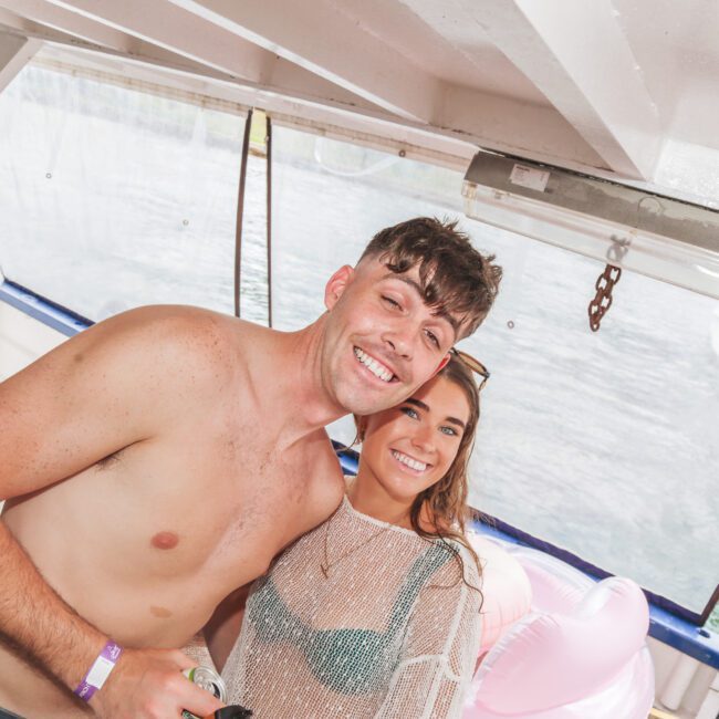 A smiling shirtless man and a woman in a mesh top and bikini pose together on a boat, with water and an inflatable pink float visible in the background.