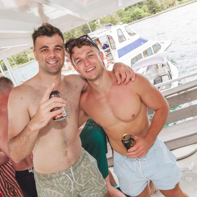 Two smiling men in swim trunks holding drinks pose arm-in-arm on a boat. Other people are socializing nearby, and several boats can be seen on the water in the background. It appears to be a sunny day.