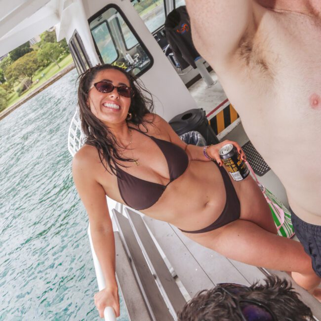 A woman in a dark bikini and sunglasses smiles and poses on a boat, holding a can drink. Two men, one shirtless, are nearby. The boat is on clear blue water with land and trees in the background.
