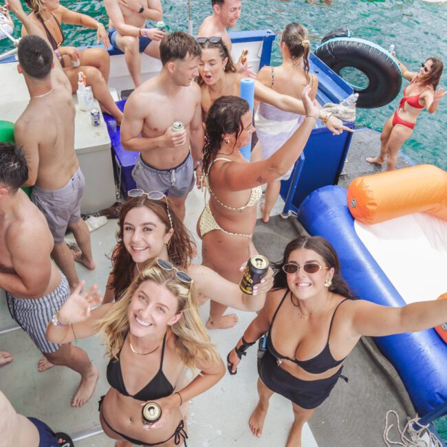 A group of young adults in swimsuits enjoy a lively boat party, holding drinks, chatting, and dancing. Inflatable floats and blue-green water surround the boat on a sunny day.