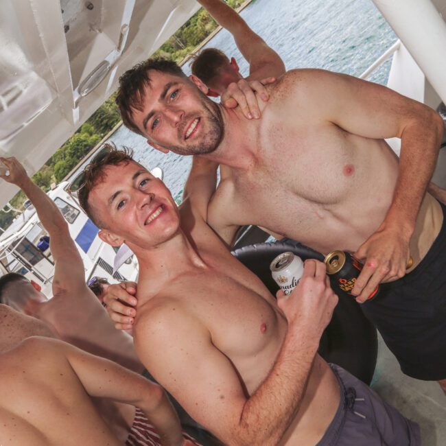 Two shirtless men smiling and posing together on a boat, each holding a drink. Other people are nearby, and water and greenery are visible in the background. The atmosphere appears casual and festive.