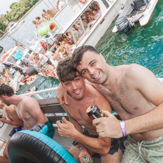 Two smiling men in swim trunks sit on the edge of a boat, holding drinks at a lively boat party. Other people relax and socialize on nearby boats, with clear blue-green water in the background.