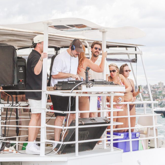 A group of people enjoying a boat party; a DJ is playing music while others dance or socialize on the deck, with water and yachts in the background on a partly cloudy day.
