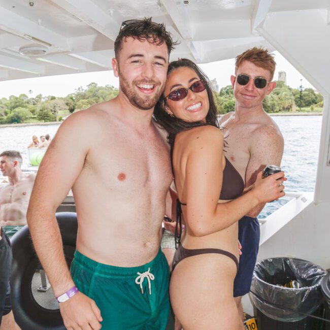 Three people in swimwear smile and pose together on a boat, with a scenic water and tree-filled background. Other people are visible seated in the background, enjoying the sunny outing.