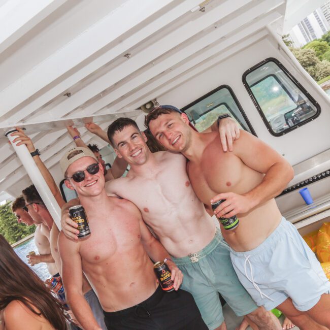 Three young men in swim trunks stand smiling and arm-in-arm on a boat, holding cans. Other people are nearby, and trees and buildings are visible in the background over the water.