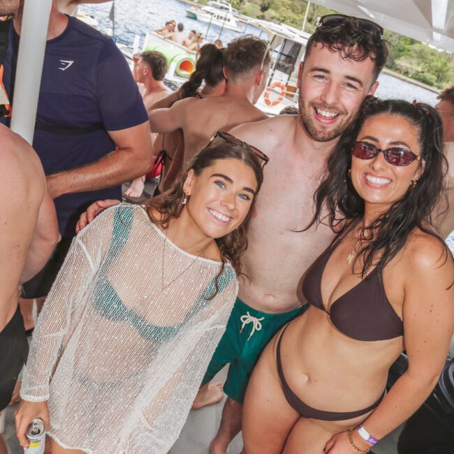 Three people in swimwear smile and pose together on a boat crowded with others. The background shows water and greenery. Two hold drinks, and the atmosphere is cheerful and relaxed, suggesting a summer boat party.