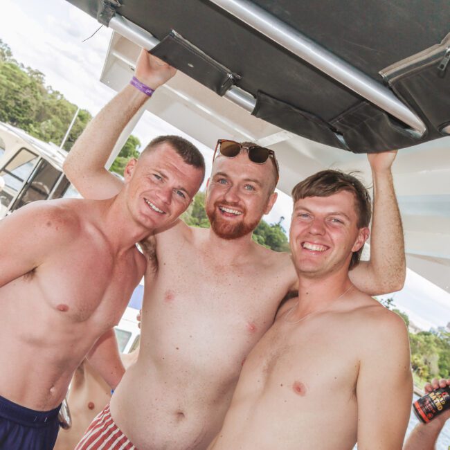 Three smiling, shirtless men pose together on a boat under a canopy, with water and trees in the background. One man wears sunglasses on his head and striped swim trunks. It appears to be a sunny day.