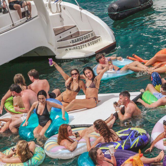 A group of people in swimsuits relax and have fun on colorful inflatables in the water near a boat, with some raising drinks and smiling at the camera on a sunny day.
