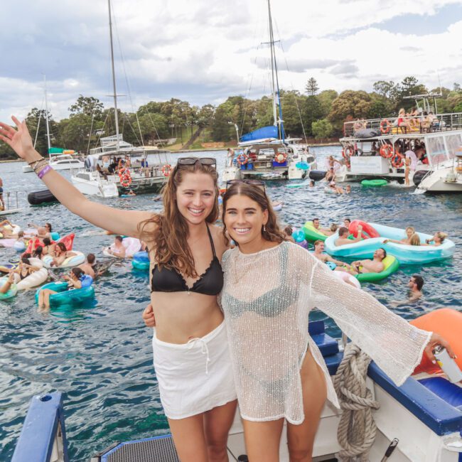 Two women in swimwear smile on a boat, surrounded by people floating on inflatables and swimming in the water. Several boats are anchored nearby, and the atmosphere is festive under a cloudy sky.