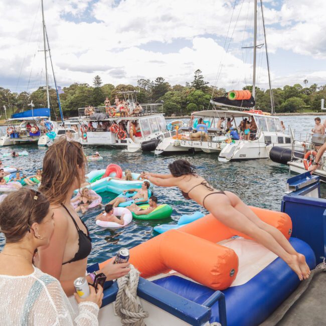 People enjoy a lively boat party on the water, with many swimming and lounging on colorful inflatables. A woman in a bikini dives from a boat as others watch, surrounded by boats and a festive atmosphere under a partly cloudy sky.