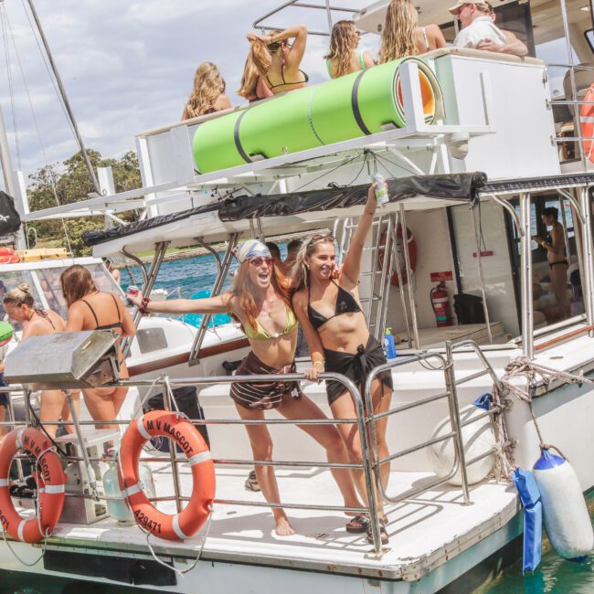 Two women in swimsuits smile and pose on the deck of a boat, surrounded by other people enjoying a lively party. Inflatable floats and safety rings are seen on the boat, with water visible in the background.