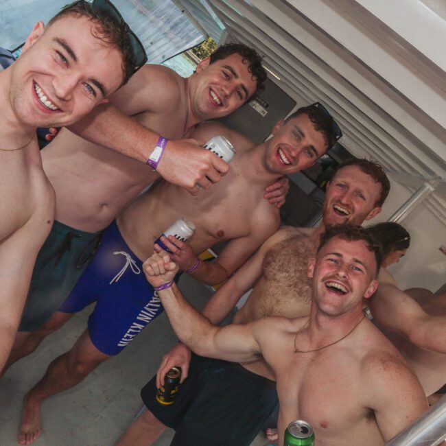 Five young men in swim shorts smile and pose together on a boat, some holding drinks, appearing to enjoy a casual, festive atmosphere.