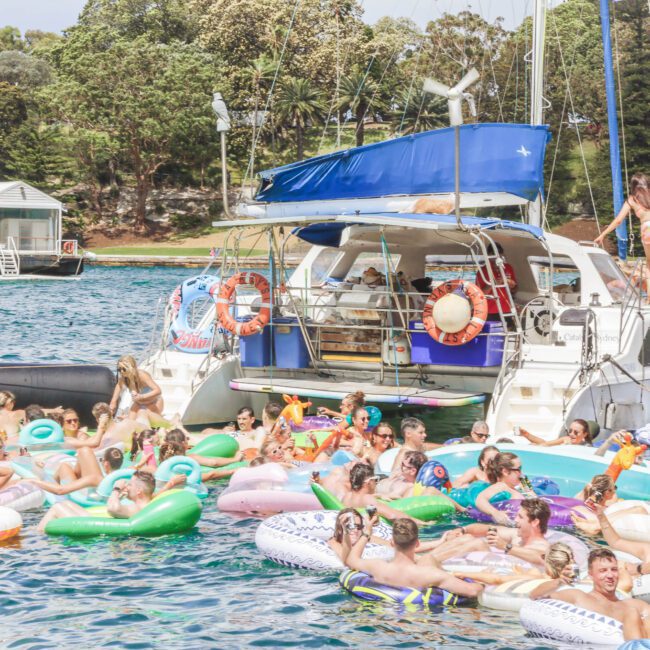 A large group of people float on colorful inflatables in the water near a docked boat, enjoying a sunny day. Trees and other boats are visible in the background.