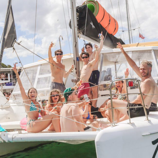 A group of smiling young adults in swimsuits sit and stand on a white sailboat, raising their arms and drinks, celebrating on a sunny day with blue skies and water in the background.