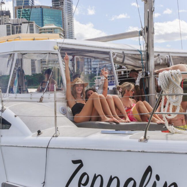 A group of people relax and smile on the deck of a white boat named Zeppelin, with city buildings and trees in the background on a sunny day. Two women in swimsuits wave and pose for the camera.