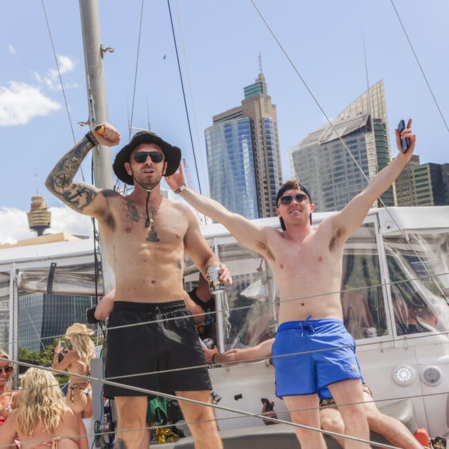 Two shirtless men in swim trunks stand on a boat, smiling and raising their arms. Other people relax in the background. Skyscrapers and a blue sky with clouds are visible behind them.