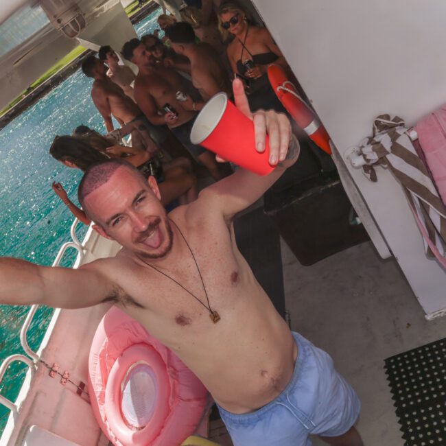 A man in swim trunks smiles and raises his arms, holding a red cup, on a boat with other people in swimwear behind him. The boat is on clear blue water and has towels and an inflatable ring nearby.