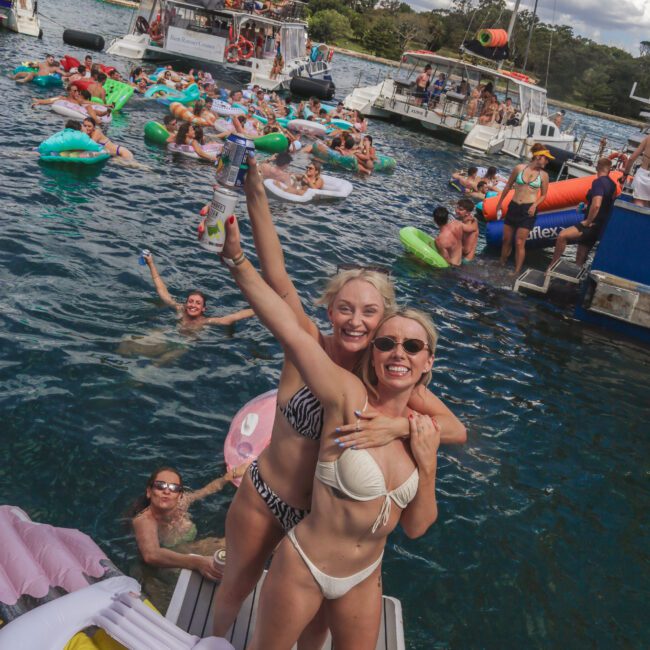 Two women in swimsuits smile and pose on a dock, one holding a drink. Behind them, people float on inflatables and swim near anchored boats on a sunny day, enjoying a lively party on the water.