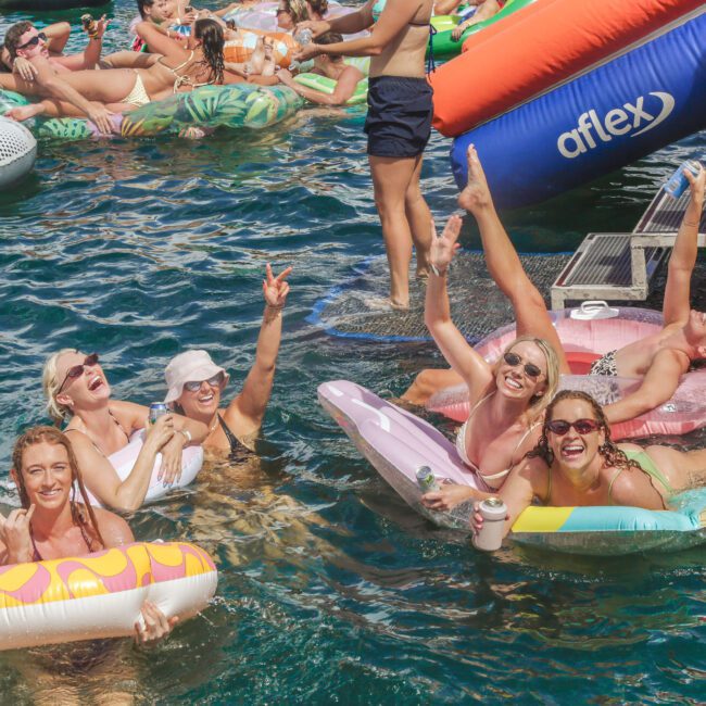 A group of smiling women in swimsuits float on colorful inflatables and pose playfully in lively, sunlit blue water during a crowded summer pool party. Others enjoy the water and inflatables in the background.