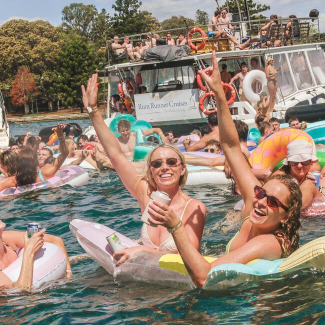 A group of people relax and have fun on colorful inflatables in the water near boats, enjoying a lively summer party. Many are smiling, raising their arms, and wearing sunglasses under a sunny sky.