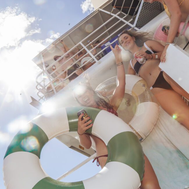 A group of young women in swimsuits smile and pose on a boat under bright sunlight, holding drinks and a green-and-white inflatable ring, with blue sky and white clouds in the background.