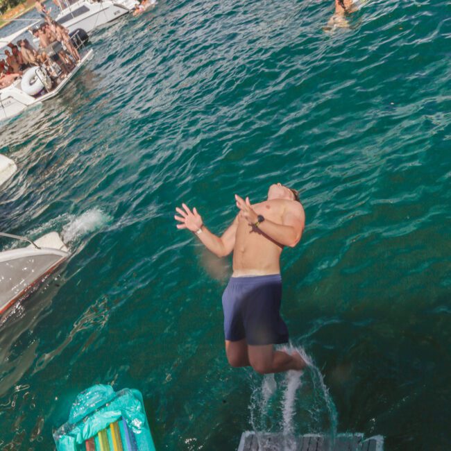 A man in swim trunks is falling backward off a boat into a lake. Several people are lounging on boats and inflatables nearby, enjoying the sunny day. The water is vibrant blue-green and filled with activity.