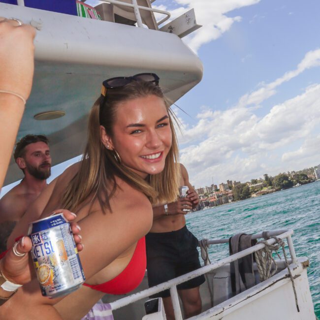 A woman in a red swimsuit smiles and leans over the railing of a boat on the water, while others enjoy the sunny day. One person holds a can of drink in the foreground. The sky is partly cloudy.