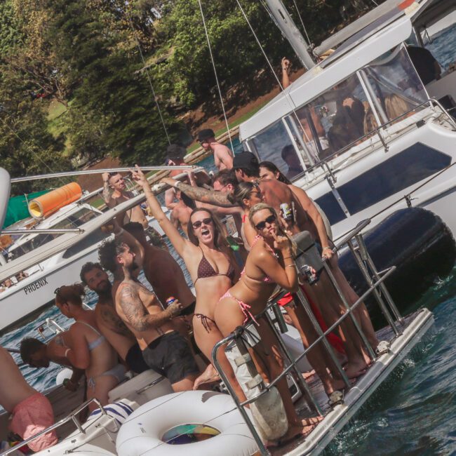 A group of people in swimsuits are partying and posing for photos on a crowded boat under the sun, with trees and another boat visible in the background.