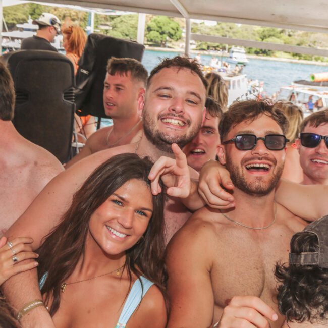 A group of young adults, mostly shirtless men, smile and pose closely together on a crowded boat during a sunny day, with water and other boats visible in the background.