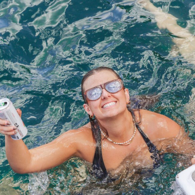 A smiling woman wearing sunglasses and a black swimsuit holds up a can while swimming in clear blue-green water, with another person partially visible beside her.